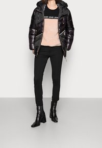 Giacca a vento nera lucida con cappuccio, abbinata a un maglione rosa chiaro in pelliccia e pantaloni skinny neri, completata da stivaletti neri con tacco.