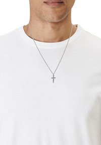 Collier en chaîne argentée avec un pendentif croix minimaliste. Le pendentif présente des détails gravés et pend sur un t-shirt blanc uni.