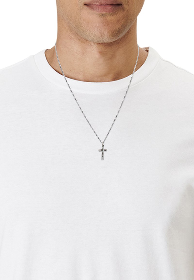 Collier en chaîne argentée avec un pendentif croix minimaliste. Le pendentif présente des détails gravés et pend sur un t-shirt blanc uni.
