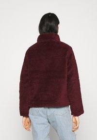 Veste polaire bordeaux avec un col montant et un tissu texturé. Présente une coupe décontractée et des détails de coutures visibles, portée avec un jean bleu clair.