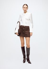 White button-up shirt with raised collar, brown suede skirt featuring gold buttons, and knee-high glossy brown heeled boots.