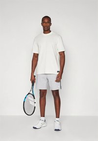 Hvid kortærmet T-shirt kombineret med grå shorts, holding en tennisracket. Har et enkelt design, blød tekstur og sporty udseende.