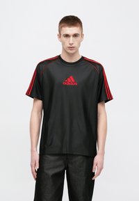 Czarna sportowa koszulka z czerwonymi akcentami, z krótkimi rękawami i okrągłym dekoltem. Na przodzie widoczne czerwone logo adidas.