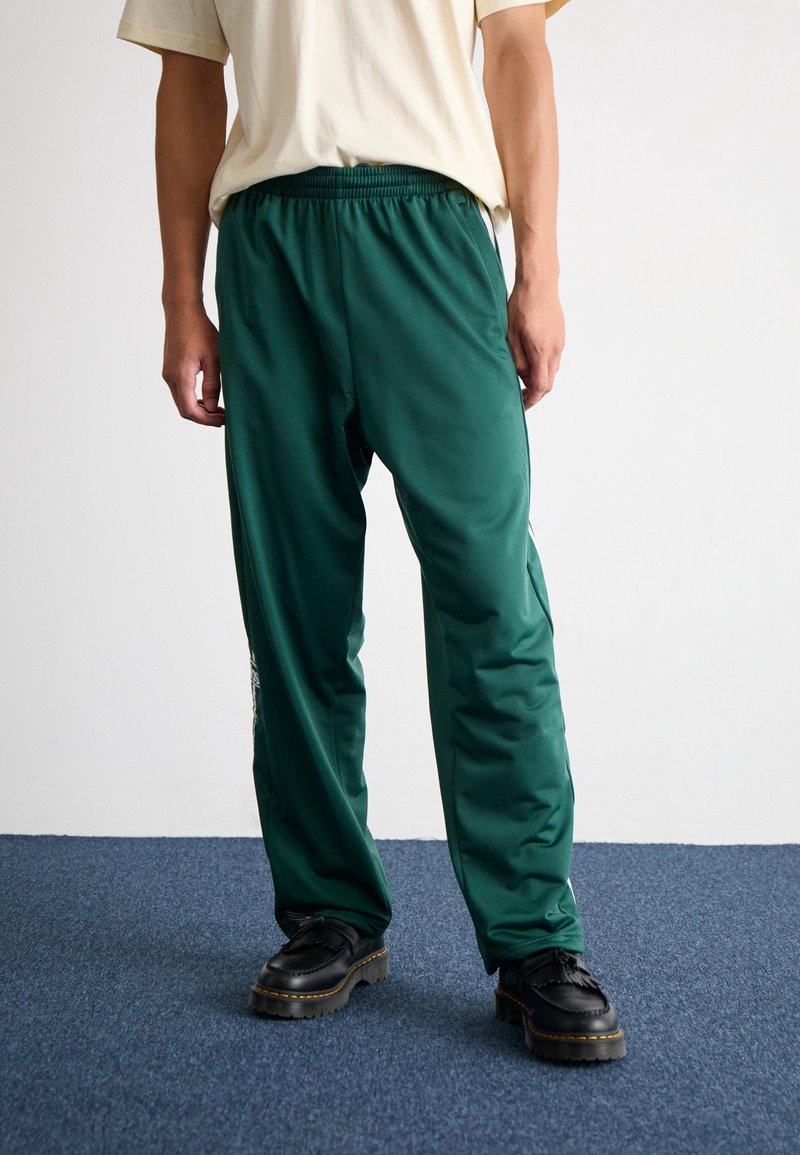 Green, lightweight athletic pants with an elastic waistband and side stripes, paired with a beige t-shirt and black lace-up shoes.