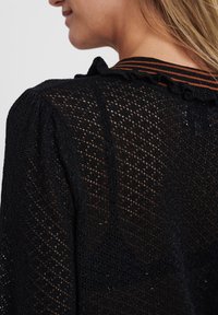 Nainen, joka käyttää mustaa läpikuultavaa neulottua yläosaa, jossa on timanttikuvioita ja liehulistakaulus, jossa on oransseja ja mustia raitoja.