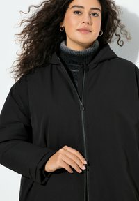 Sort lynlåses jakke med hætte i tekstureret stof, elastiske manchetter og høj krave med en grå striktrøje under.