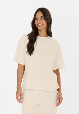 Lachende vrouw met lang donker haar die een loszittend beige shirt met korte mouwen en bijpassende broek draagt, staand tegen een witte achtergrond.