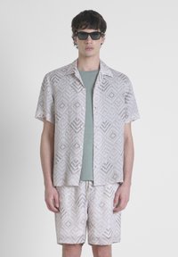 Completo con camicia a maniche corte e pantaloni corti in beige chiaro con un motivo geometrico, abbinato a una t-shirt teal e occhiali da sole neri.