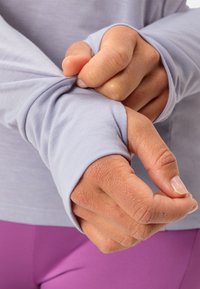 Maglietta a maniche lunghe, azzurra chiara con polsini aderenti. Mani che tirano le maniche, mostrando la texture morbida del tessuto e pantaloni leggeri viola sullo sfondo.