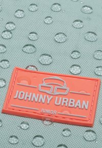 Oranžová gumená štítka s textom "JOHNNY URBAN JUNIOR", ležiaca na textúrovanej mätovej látke s vodnými kvapkami.