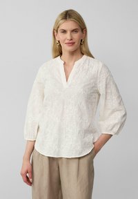 Witte geborduurde blouse met een V-hals en driekwart mouwen, voorzien van bloemmotieven en een zachte, gestructureerde stof, gecombineerd met beige broeken.