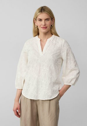 Blouse blanche brodée avec un décolleté en V et des manches trois-quarts, ornée de motifs floraux et d'un tissu doux et texturé, associée à un pantalon beige.