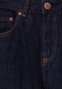 Gros plan sur un jean en denim bleu foncé montrant la poche avant, les rivets en cuivre, la couture orange et la fermeture par bouton en cuivre.