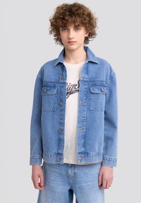 Giacca di jeans azzurro chiaro con chiusura a bottoni, due taschini sul petto, vestibilità rilassata e cuciture in contrasto, indossata sopra una maglietta bianca.