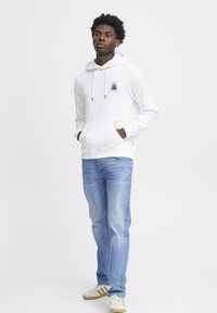 Sweat à capuche blanc avec cordon de serrage ajustable, poche avant et détail de logo. Associé à un jean bleu clair et des baskets crème avec des rayures.