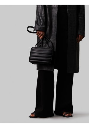 Personne portant un long manteau noir texturé et un large pantalon noir, tenant un petit sac à main noir matelassé, chaussée de sandales noires ouvertes.