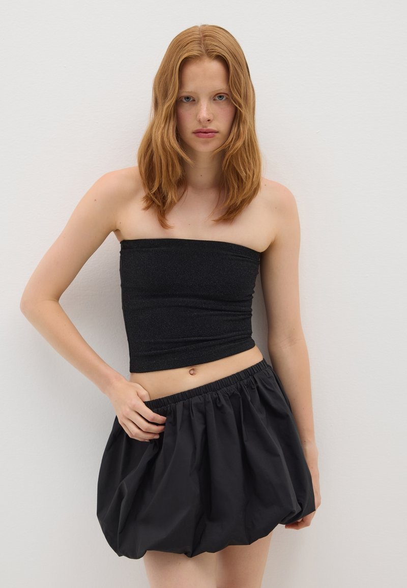 Giovane donna con capelli rossi che indossa un top nero senza spalline e una gonna nera voluminosa, in piedi contro un muro bianco semplice.