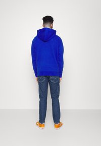 Sudadera con capucha azul de textura suave, combinada con jeans de mezclilla oscuros que tienen bolsillos laterales y zapatillas naranjas con detalles en blanco.