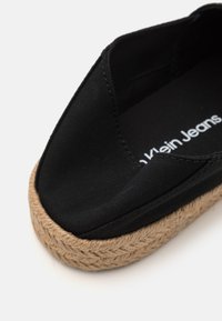 Chaussure slip-on en toile noire avec un bout fermé, dotée d'une semelle extérieure en corde de jute texturée et d'un intérieure en tissu doux. Logo visible à l'intérieur.