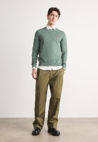 Light green knit sweater layered over a white collared shirt, paired with loose olive-green trousers and black shoes.