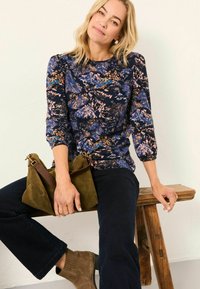 Bluse mit Blumenmuster in Marineblau, Pink und Blau, mit Dreiviertelärmeln. Dunkelblaue, ausladende Jeans und eine olivgrüne Handtasche. Wildleder-Stiefeletten.