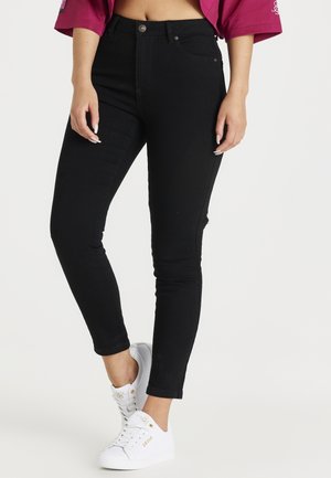 Frau trägt schwarze Skinny-Jeans mit hoher Taille und weiße Sneakers, die Hände entspannt an den Seiten und ein kurzes magentafarbenes Oberteil.