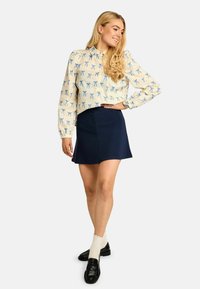 Kremna bluza z modrimi vzorci pentelj, mornarsko modro mini krilo, kremne rebraste nogavice in črni mokasini. Gladek material z urejeno obliko.