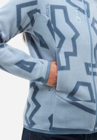 Hellblaues Fleece-Jacke mit dunkelblauen geometrischen Mustern, einer Reißverschlusstasche an der Seite und einer Hand, die teilweise in der Tasche ist.