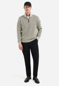 Man in een grijze gestructureerde sweater met kwartrits, zwarte broek, zwarte pet en zwarte laarzen, staand met één hand in zijn zak op een witte achtergrond.