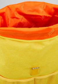 Bolsa de tela amarilla con una parte superior de nylon naranja con cordón, que presenta un acento metálico y un fondo de malla para ventilación.