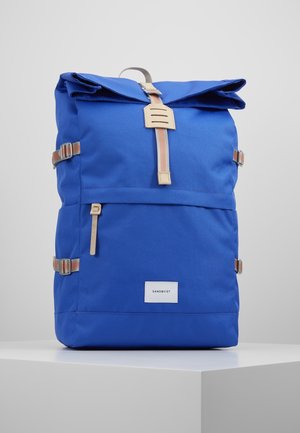 Sac à dos bleu à fermeture roulante avec poche avant, bretelles rayées beige et orange, et étiquette Sandqvist sur une surface blanche.