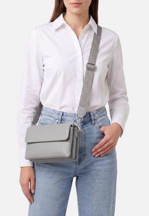 Donna che indossa una camicia bianca con bottoni e jeans azzurri chiaro che tiene in mano una borsa a tracolla grigio chiaro con tracolla larga e regolabile.