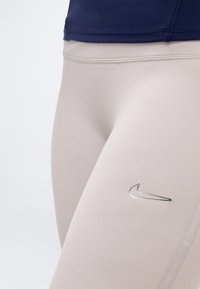 Κοντινό πλάνο σε αθλητικό κολάν απαλού μπεζ χρώματος με ασημί λογότυπο Nike στο μηρό και ορατό σκούρο μπλε επάνω άκρο.