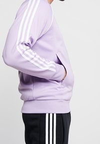 Lila sweatshirt med långa vita ränder på ärmarna, med en sidoficka och ribbade muddar, ihop parat med svarta byxor med liknande ränder.