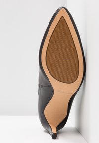 Zapato de tacón alto negro de cuero con punta afilada. Suela de goma texturizada en color marrón claro, con un diseño curvado que presenta una marca en relieve.