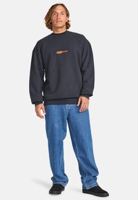 Sudadera oscura de color azul marino con cuello redondo y logo naranja. Combinada con jeans holgados de color azul claro y zapatos negros. Ajuste cómodo y casual.