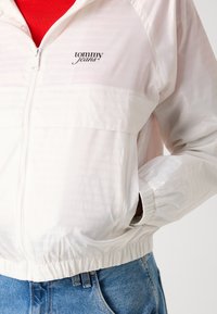 White windbreaker jacket with repeating "tommy jeans" pattern, elastic cuffs and hem, zip closure, and a chest logo detail.