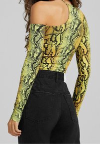 Långärmad, åtsittande topp med ett gult och svart ormtryck, med en öppen axeldesign, kombinerad med svarta högmidjadejeans.