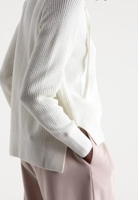 Gebreide trui in off-white, met een geribbelde textuur, lange mouwen, zijslits en een losse pasvorm, gedragen met lichtroze broek.