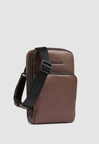 Borsa a tracolla in pelle marrone con finitura testurizzata, tracolla nera regolabile, scomparto con zip singola e logo impresso sul davanti.