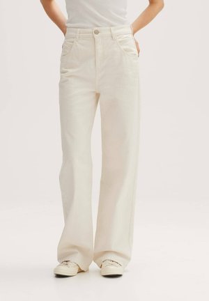 Person wearing high-waist wide-leg cream-colored pants with pockets and off-white sneakers, standing with hands in pockets.