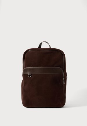 Mochila de ante marrón oscuro con bolsillo delantero con cremallera, detalles en cuero, anilla metálica y asa superior para llevar, sobre un fondo neutro.