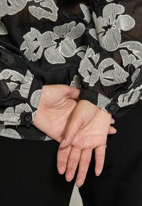Crna bluzica s bijelim cvjetnim vezom, prozirna tkanina i manžete s gumbima. Ruke su spojene iza leđa, a dugi nokti su vidljivi.