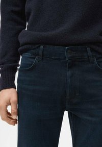 Vaqueros de mezclilla de color azul oscuro, con un diseño estándar de cinco bolsillos, cierre de botón y corte slim. Acompañados por un suéter oscuro.