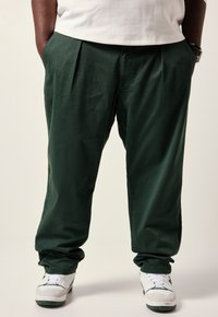 Groene geplooide broek van een lichte stof, met een relaxed fit. Draagt witte sneakers met groene accenten.