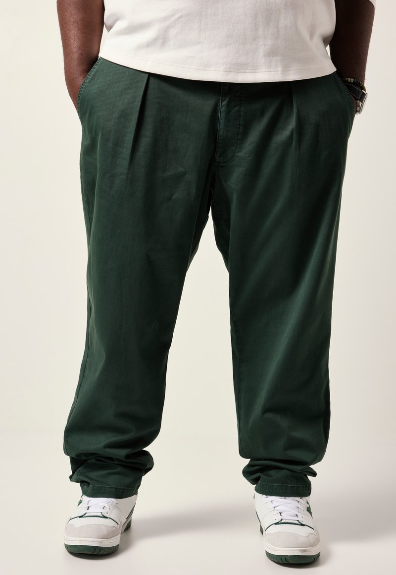 Groene geplooide broek van een lichte stof, met een relaxed fit. Draagt witte sneakers met groene accenten.