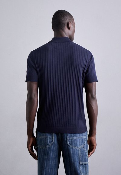 Polo shirt côtelée marine à manches courtes, fabriqué à partir d'un matériau texturé. Présente un col et un design ajusté, associé à un pantalon en denim bleu.