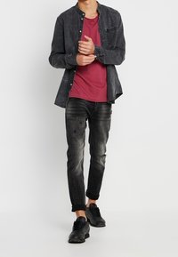 Camicia nera in denim a maniche lunghe sopra una t-shirt a collo rotondo color bordeaux. Indossata con jeans neri strappati e sneakers nere. Design casual e aderente.