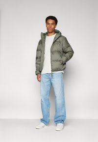 Chaqueta acolchada verde oliva con capucha, cierre de cremallera y dos bolsillos frontales, usada sobre una camiseta gris clara y unos jeans de pierna ancha azul.