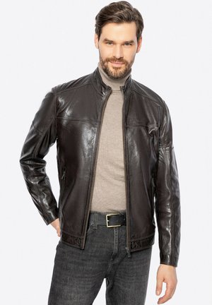 Chaqueta de cuero marrón con cremallera frontal, cuello de pie y textura suave. Llevada sobre una camiseta gris claro y combinada con jeans oscuros.